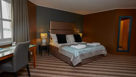 A neatly made double bed with pillows and towels on top is set in a cozy hotel room with striped carpet, a desk and chair by the window, and a large mirror on the wall.