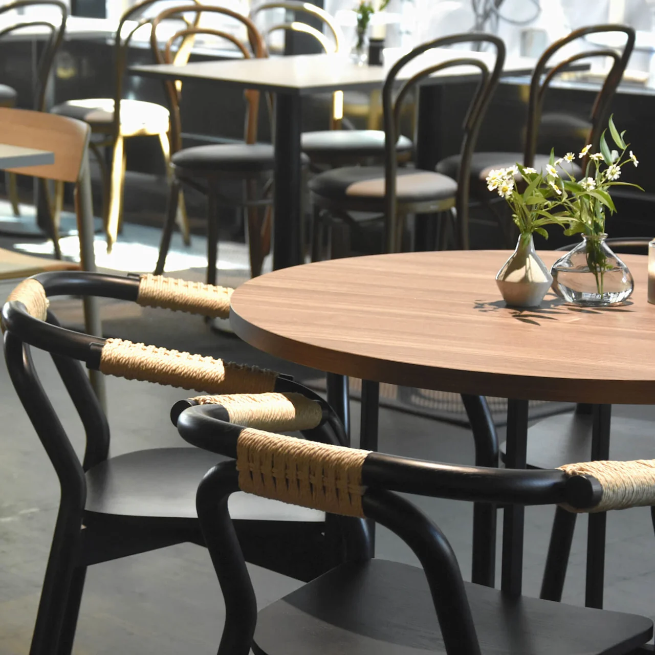 A round wooden table holds a small vase with flowers and a candle. Around it, black chairs with woven backs are placed, in a sunlit cafe setting.  