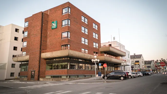 A multi-story red brick hotel building displaying a "Quality Hotels" sign, situated on a street corner, with parked cars along the sidewalk.