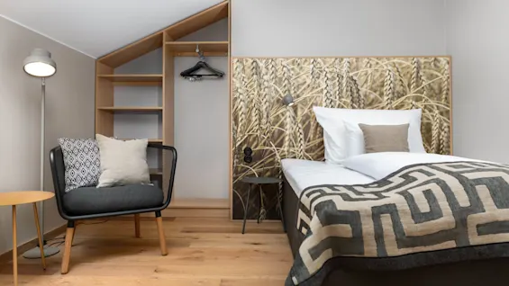 A well-lit, tidy hotel room with wooden floors, a plush bed, patterned headboard, a cozy chair, pillows, a standing lamp, and built-in shelves.