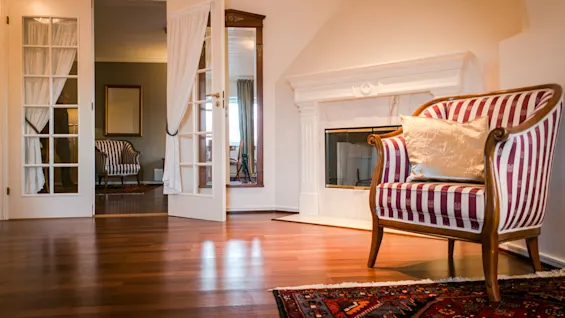 An elegant armchair with red and white stripes sits in front of a fireplace on a wooden floor, surrounded by a mirror and open French doors revealing another room.