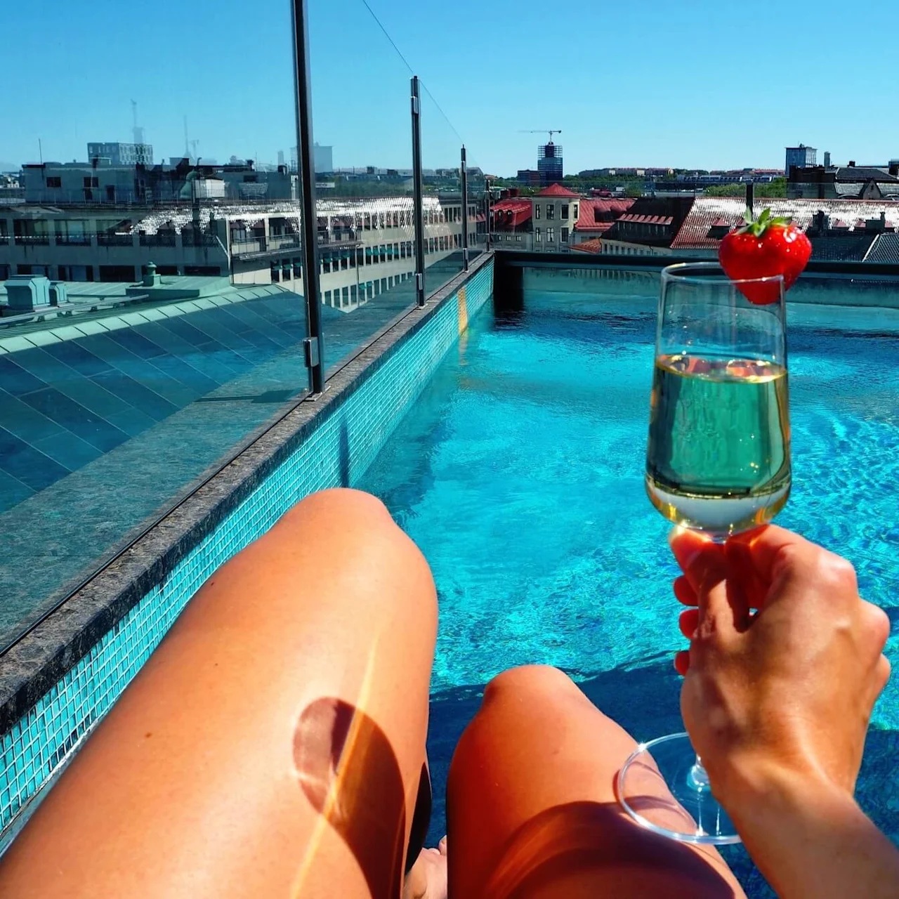 A hand holds a champagne glass with a strawberry garnish, overlooking a rooftop pool surrounded by city buildings under a clear blue sky.