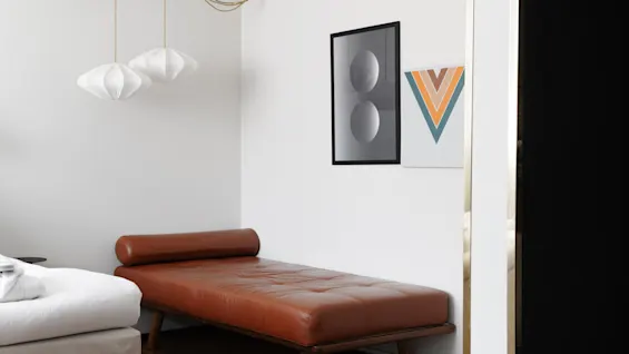 En brun lädersoffa står bredvid en säng, med geometrisk konst och hängande lampor, i ett minimalistiskt rum.