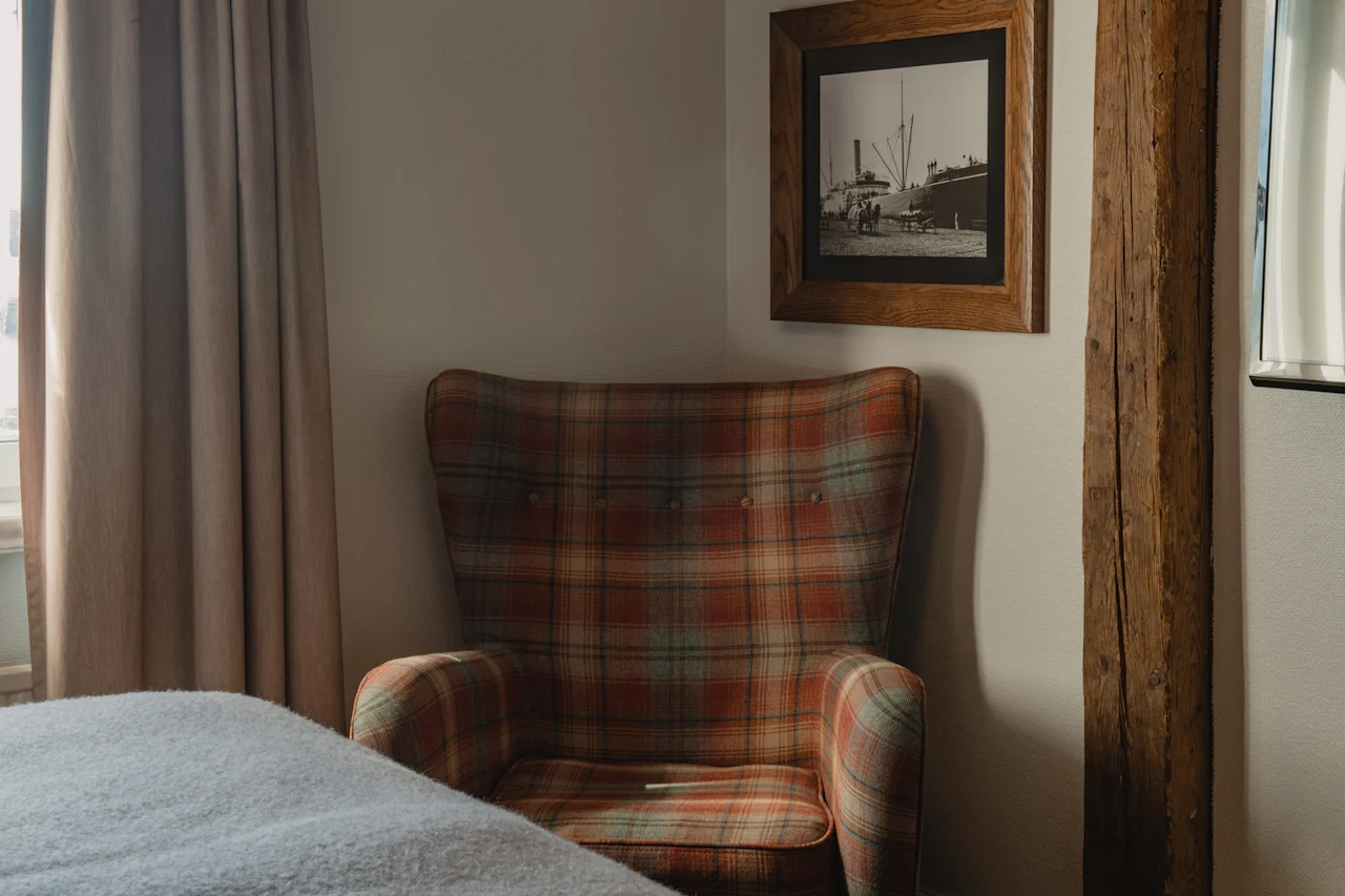 Et varmt og indbydende hjørne af et værelse med en ternet lænestol, en seng og et indrammet nautisk print.