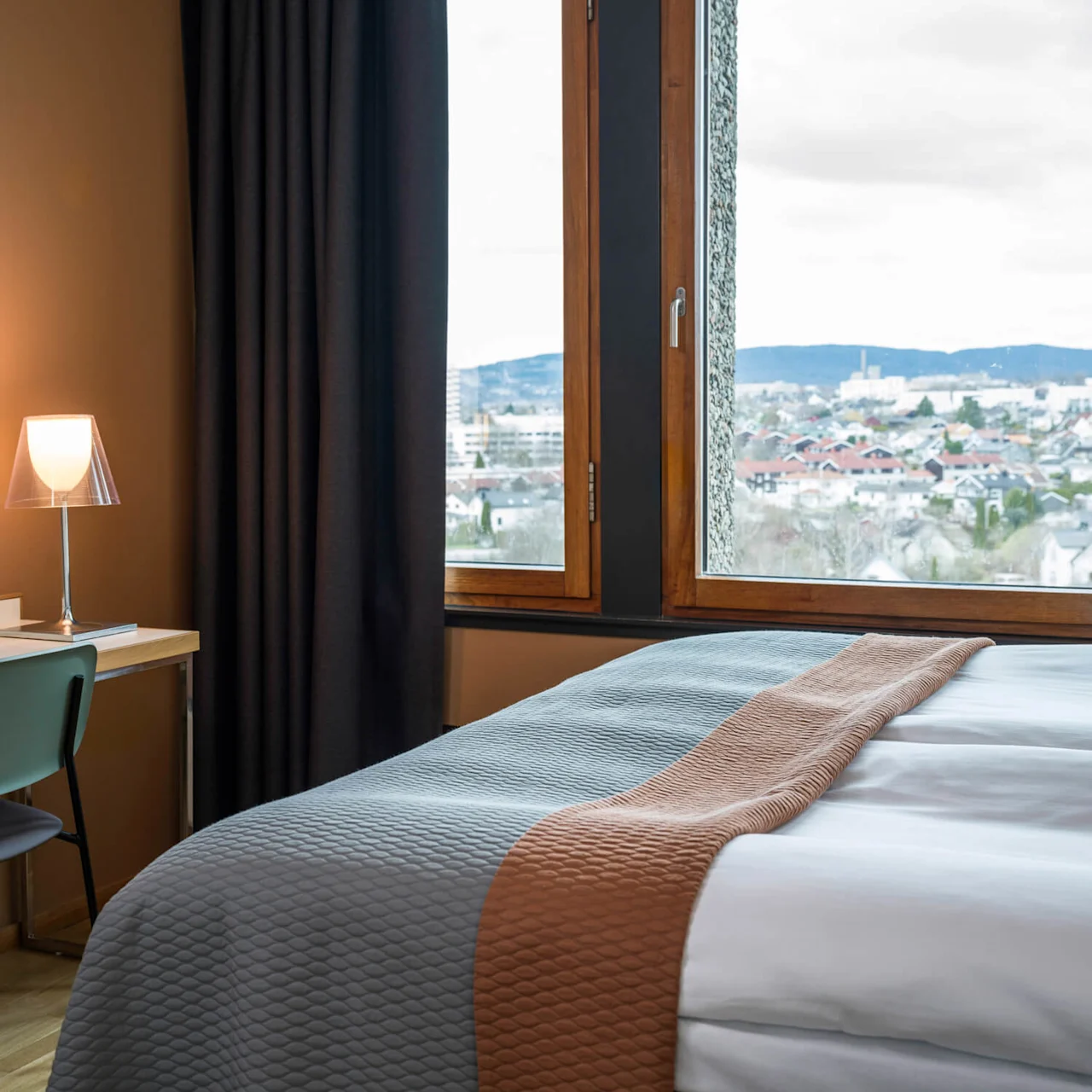 A hotel room features a bed with a gray and brown cover, a wall-mounted TV, and a desk with a lamp and chair. Large window reveals city and mountain view.