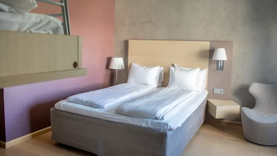 A neatly made double bed with white linens is positioned against a wall, flanked by lamps. Nearby, a light-colored chair and wooden furnishing create a cozy bedroom atmosphere.