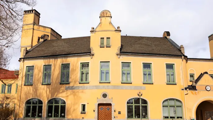 En gul bygning med grønnkantede vinduer og en mønstret tredør står i en brosteinsbelagt gårdsplass. Over inngangen leses teksten utydelig "ASCHEBERGAGYMNASIET BOLIDENHUSE." En klokke er synlig til høyre.