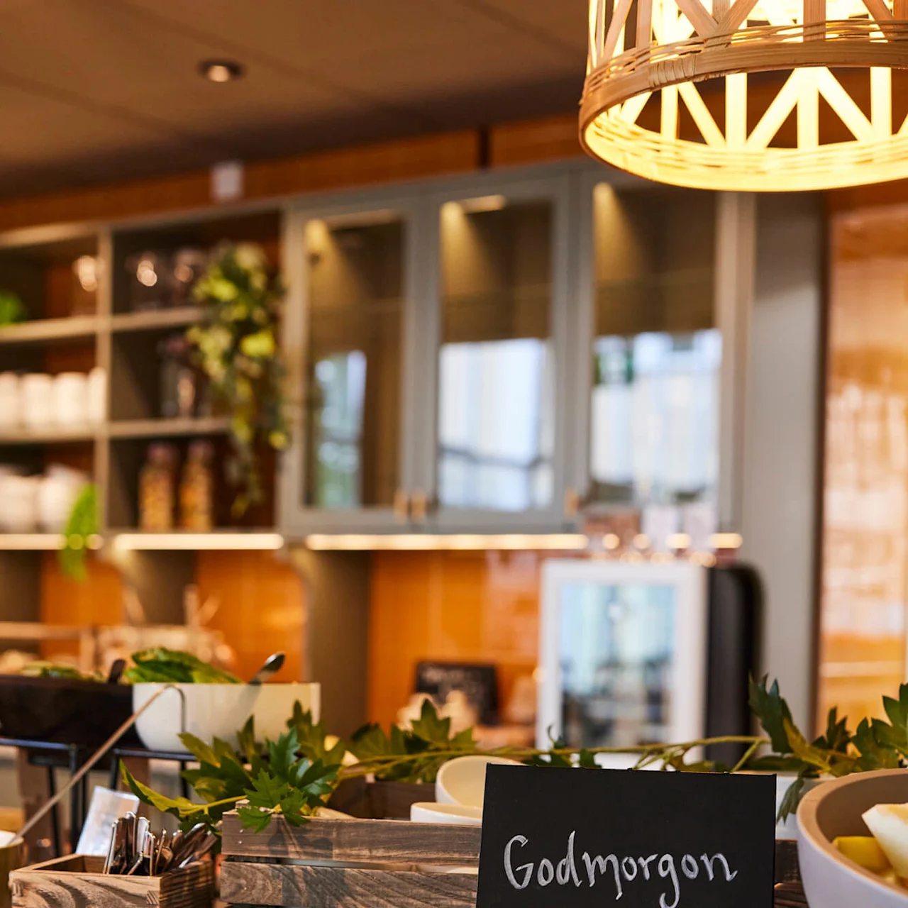 En svart tavle med "Godmorgon" og et hjerte er plassert på en kjøkkenbenk omgitt av planter og trekasser, under et varmt, dekorativt lys.
