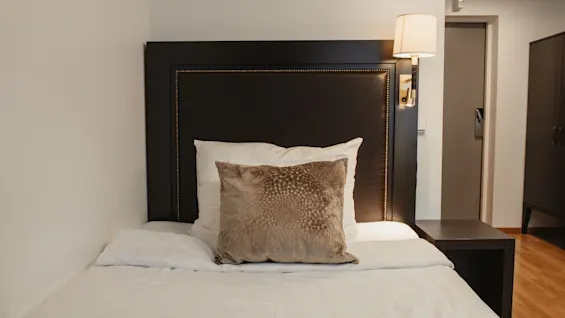 Viihtyisä yhden hengen sänky valkoisilla liinavaatteilla ja koristetyynyllä, yöpöytävalaisimen ja tumman sängynpäädyn vieressä modernissa huoneessa.