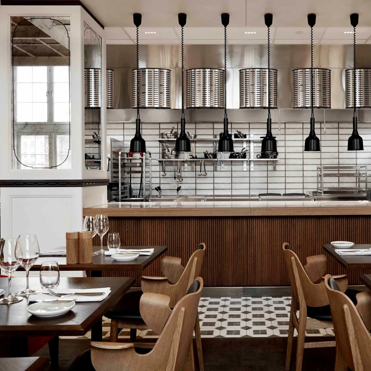Chic restaurant interior; wooden tables set with plates and glasses. Overhead, modern pendant lights illuminate an open kitchen with industrial accents and tiled walls.