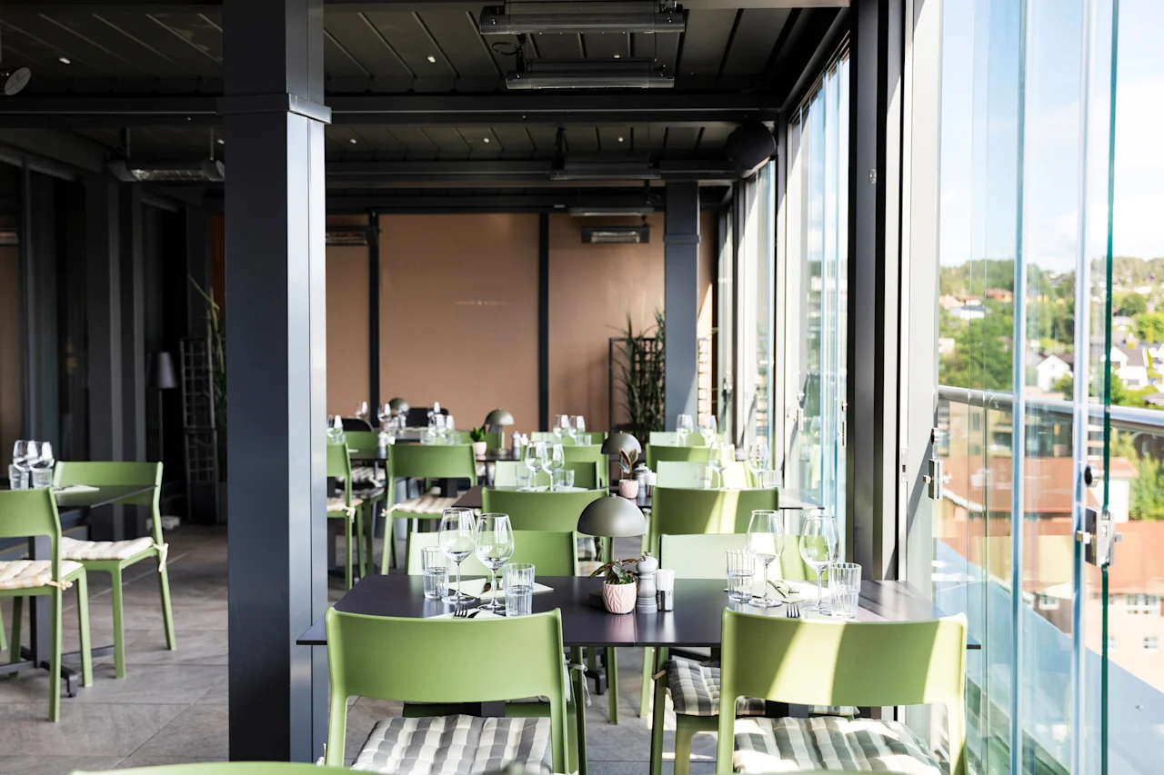En lys, moderne restaurant med grønne stoler, mørke bord og store vinduer med utsikt over en by.