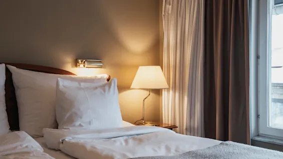 Ett mysigt hotellrum med en bäddad säng, varm belysning från en sänglampa och gardiner bredvid ett fönster.