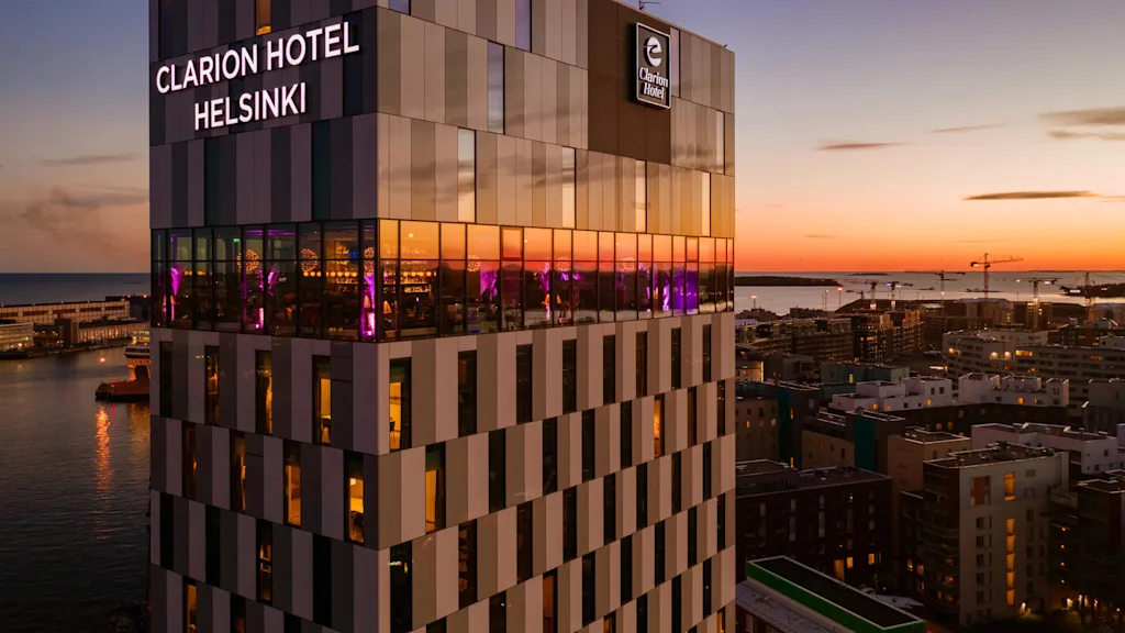 Clarion Hotel Helsinki reser sig över en hamn vid solnedgången, med reflekterande fönster upplysta av lila ljus inomhus. Omgivande stadsbild och vatten skapar en rofylld kvällsatmosfär.