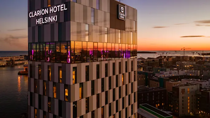 Clarion Hotel Helsinki rager over en havn ved solnedgang, med reflekterende vinduer opplyst av lilla lys innendørs. Omgivende bylandskap og vann skaper en fredelig kveld.