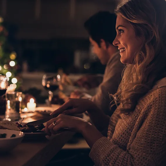En person smiler mens den spiser ved et festlig bord med et uskarpt juletre og varme lys i bakgrunnen.