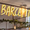 En ljusgul neonskylt med texten "BARCEPTION" hänger ovanför en hylla med gröna rankor och glas i en modern inomhusmiljö.