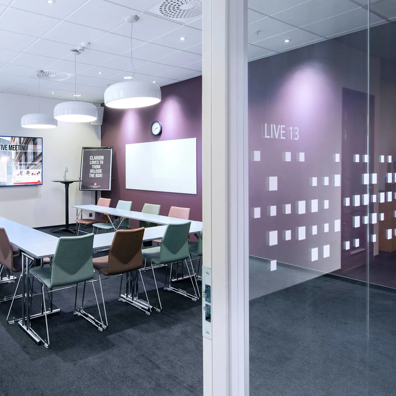 Møterom med langbord og forskjellige stoler, en stor skjerm som viser "ENJOY YOUR CREATIVE MEETING!" og en flippover med teksten "CLARION LOVES TO THINK OUTSIDE THE BOX!" Glassdører med "LIVE 13."