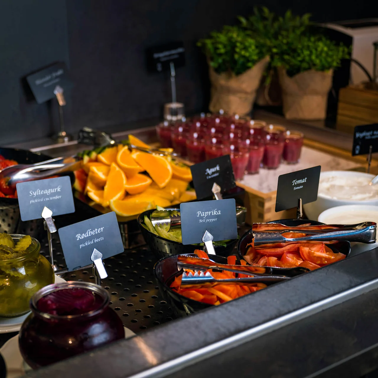 Bowls of fruits, vegetables, and condiments are arranged on a buffet table with small signs. Text includes: "Sylteagurk," "Rødbeter," "Paprika," "Tomat," and "Majones."