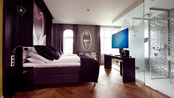 Bed with plush pillows against a swan mural; modern room features wood flooring, frosted glass shower, TV, and mirrored cabinet under soft lighting.