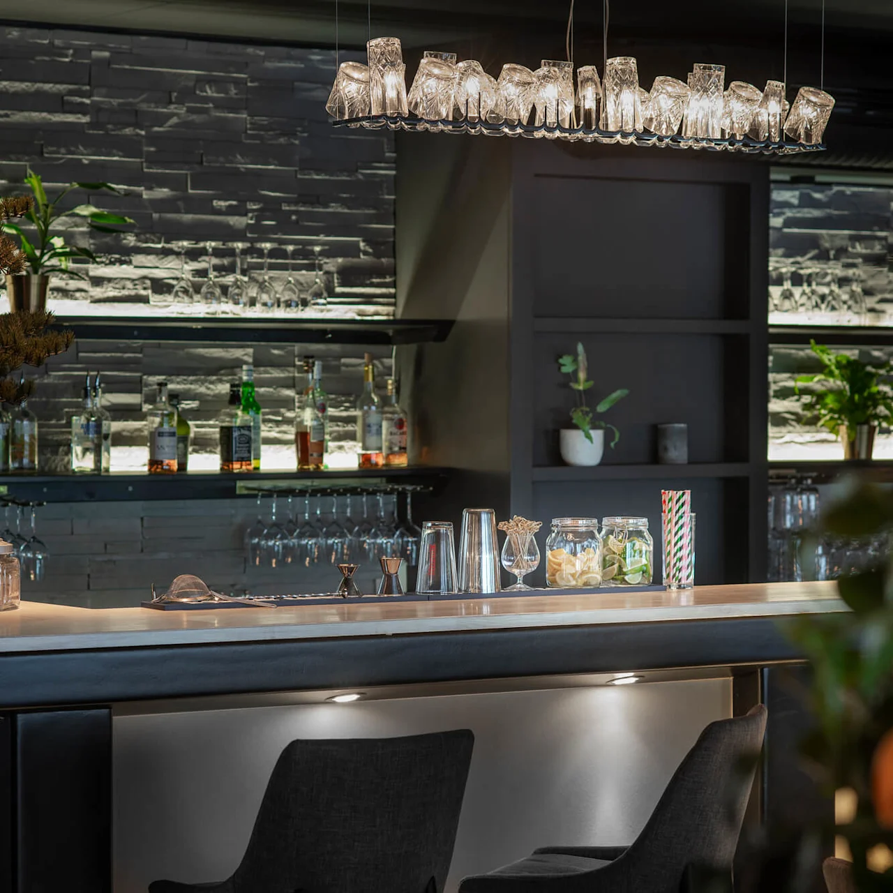 En modern bardisk med glasburkar och flaskor, med en dekorativ hängande lampa ovanför. I bakgrunden visas upplysta hyllor med olika flaskor, växter och uppochnervända glas.