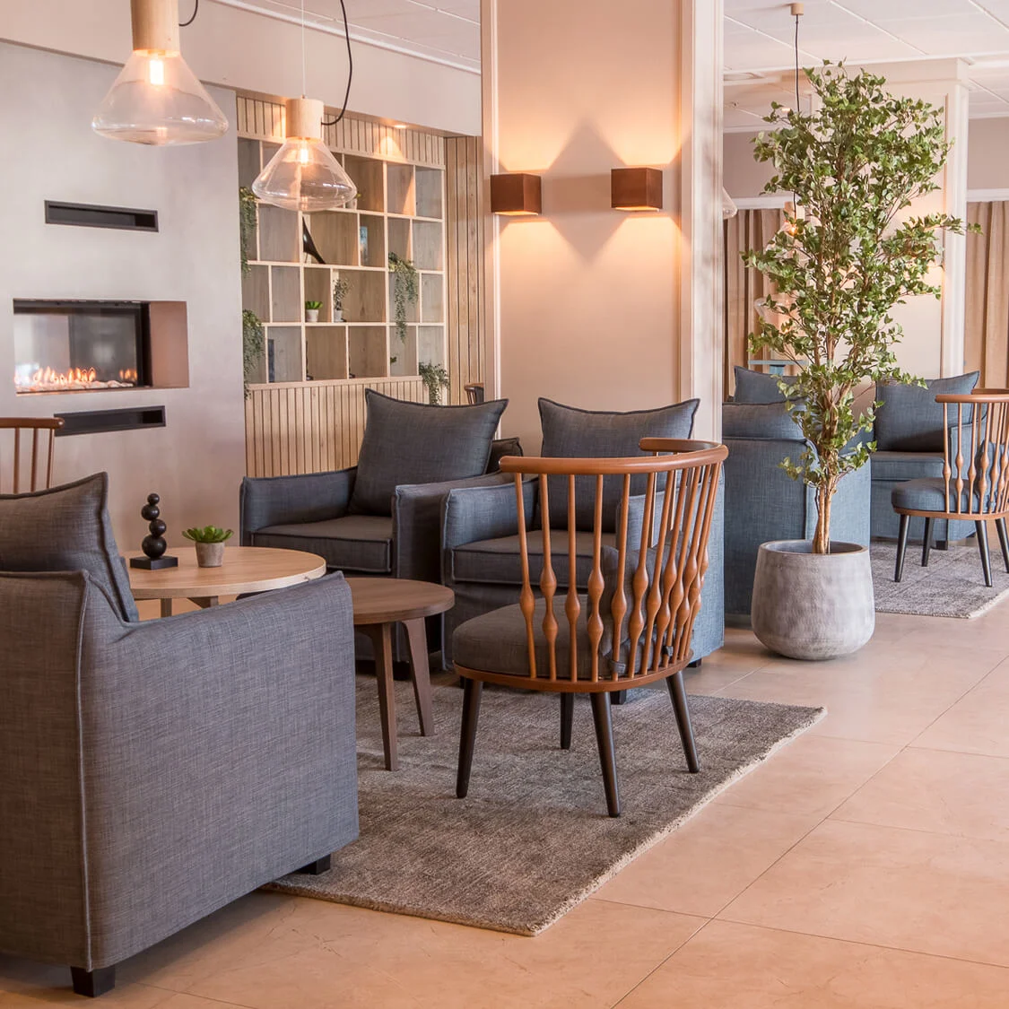 Elegant loungeområde med grå lenestoler og trestoler rundt runde bord på tepper, med en peis i bakgrunnen. Pottede planter og myk belysning forbedrer den koselige atmosfæren.