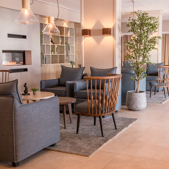 Elegant loungeområde med grå lenestoler og trestoler rundt runde bord på tepper, med en peis i bakgrunnen. Pottede planter og myk belysning forbedrer den koselige atmosfæren.