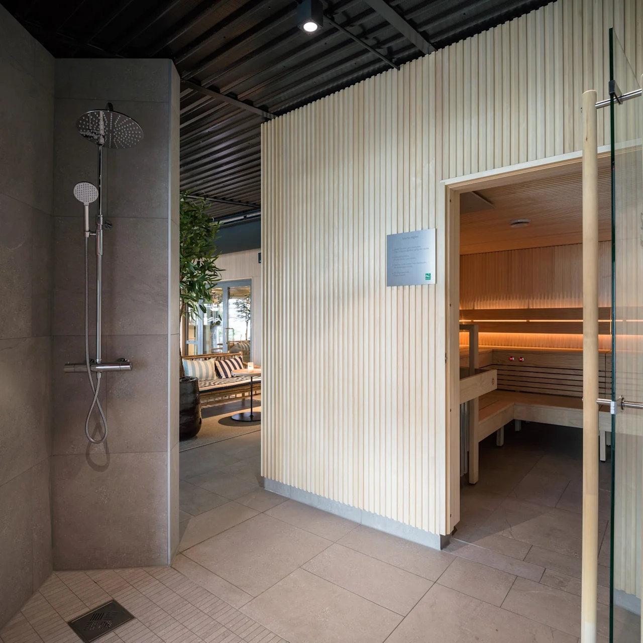 Dusch och tvålpump bredvid öppen dörr till en träbastu i en modern spa-miljö. En skylt bredvid dörren lyder: "Puulämmitteinen sauna". Bekväm lounge med växter synlig i bakgrunden.