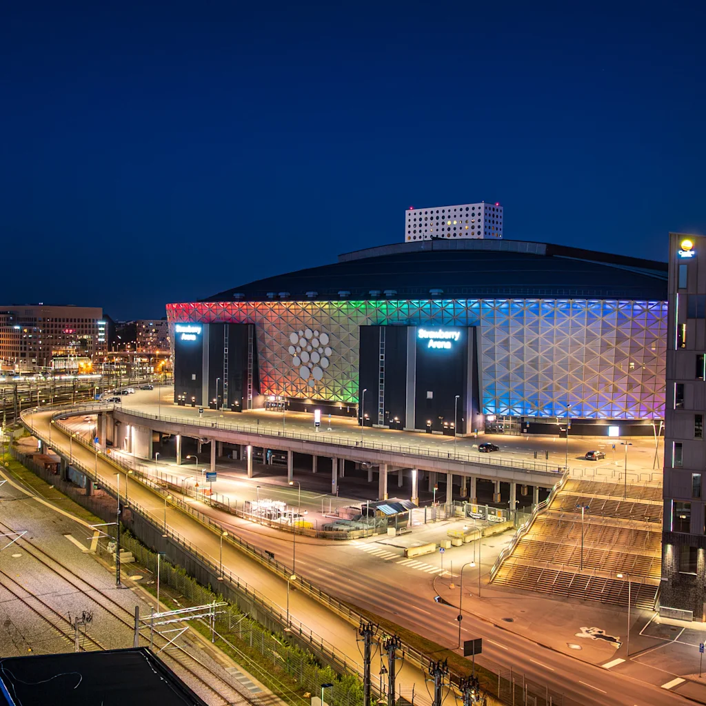 Night view of Strawberry Arena illuminated in rainbow colors for Pride, surrounded by city lights and roads.