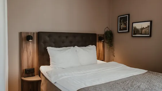 Viihtyisä hotellihuone, jossa on siististi pedattu sänky, yöpöytälamput ja kehystettyjä taideteoksia seinällä, luoden kutsuvan tunnelman.