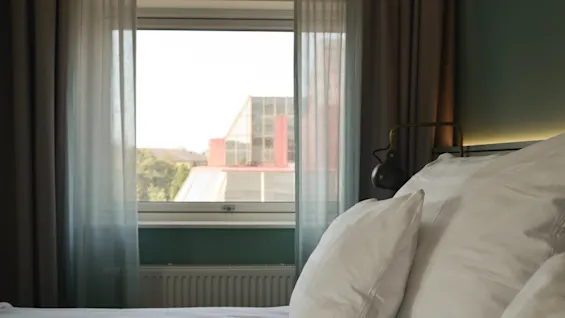 Seng med hvide puder står foran et blødt bagbelyst vindue med tynde gardiner; en bygning og grønne områder kan ses udenfor.
