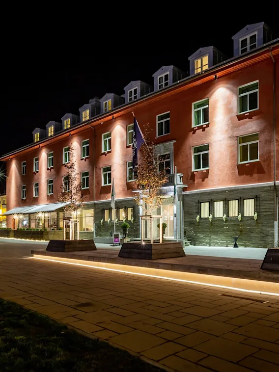 En godt opplyst bygning om natten, muligens et hotell, med trær i plantekasser og opplyste stier som fører til inngangen.