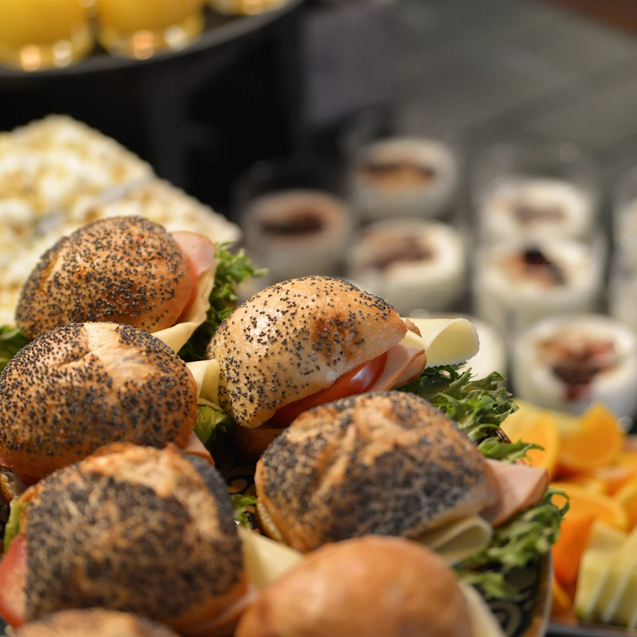 Sandwiches fyldt med kød og ost er arrangeret på et fad, pyntet med salat. I baggrunden vises yoghurtbægre og appelsinjuice på et bord.