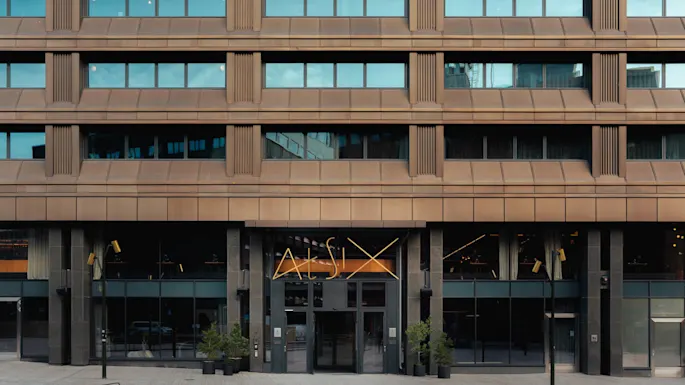 A brown multi-story building facade with large windows and a modern entrance featuring the text "At Six." Potted plants and a curved bench are positioned in the courtyard.