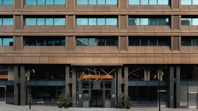A brown multi-story building facade with large windows and a modern entrance featuring the text "At Six." Potted plants and a curved bench are positioned in the courtyard.