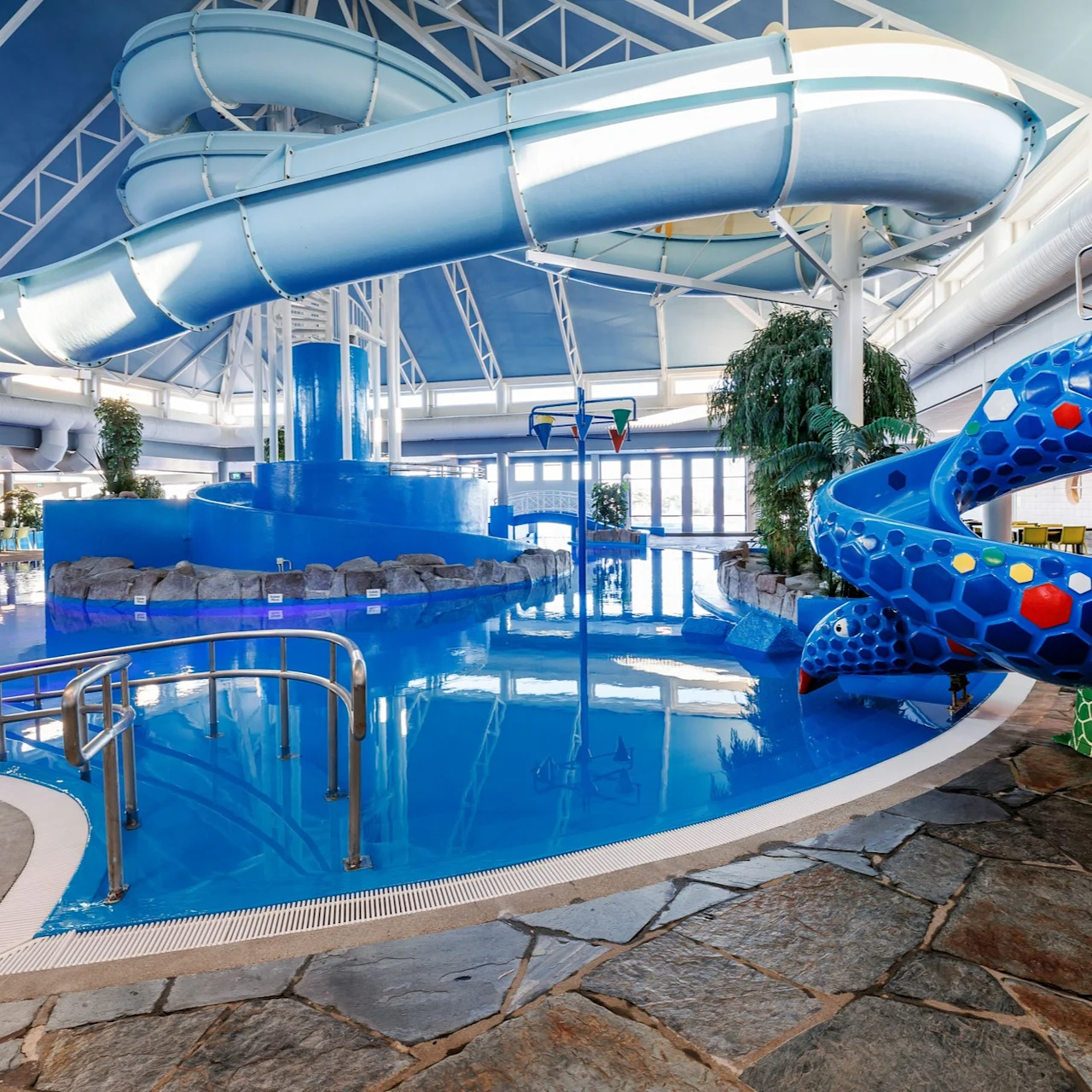 An indoor pool features two blue slides, one twisting and one tunnel-type, surrounded by stone floor and tropical plants under a glass ceiling.