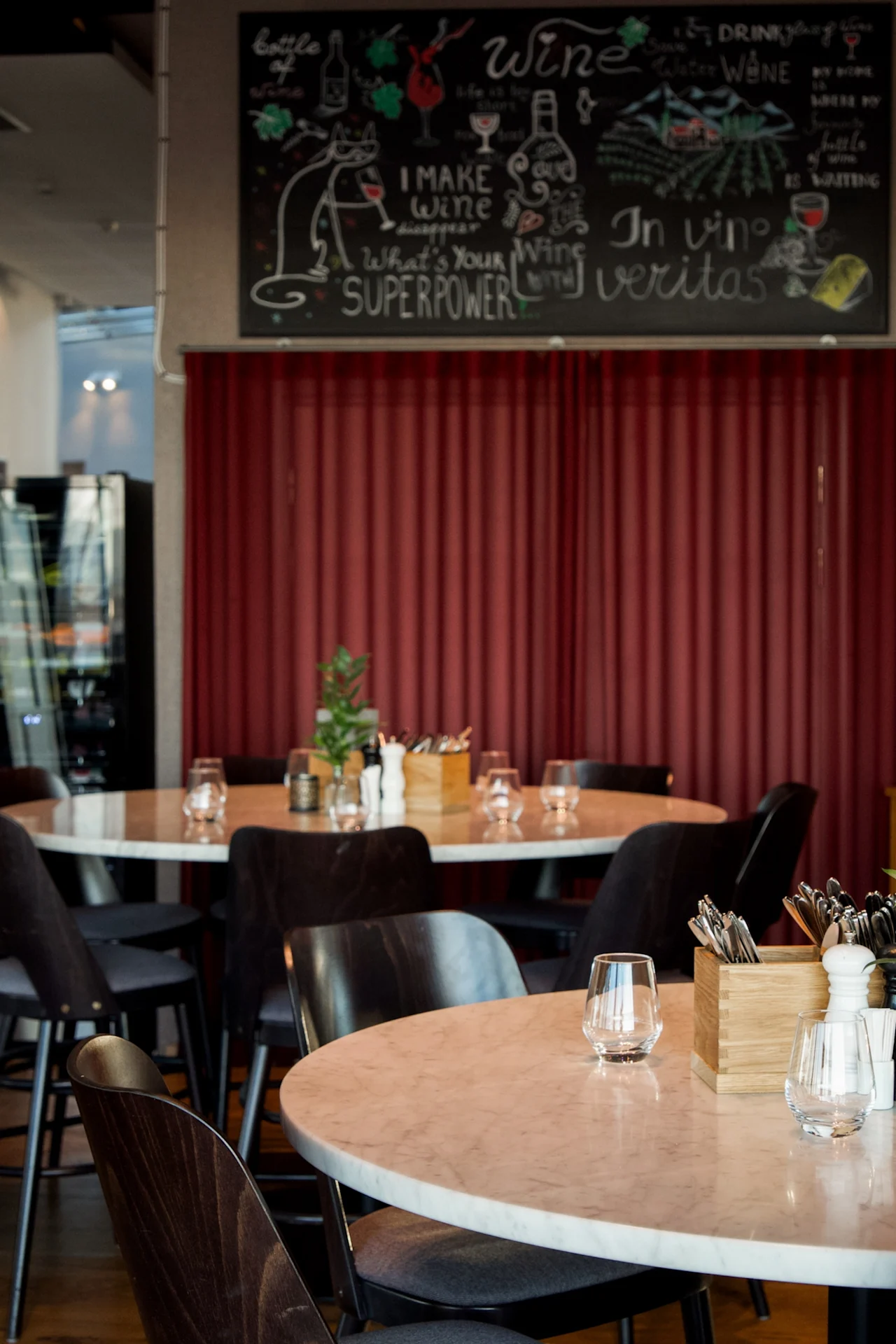 Runde bord med glass og bestikkholdere er arrangert i en restaurant. En tavle ovenfor sier: "Wine, I MAKE wine disappear, What's Your SUPERPOWER, In vino veritas," med vinrelaterte tegninger.