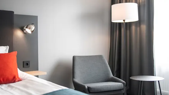 En grå fåtölj står bredvid ett litet runt bord och en golvlampa, i ett modernt rum med en säng med en röd kudde och ett fönster med mörka gardiner.