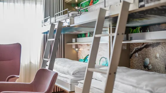 Bunk beds with white bedding and ladders, situated in a brightly lit room. A pink armchair sits nearby, with large windows allowing natural light to fill the space.