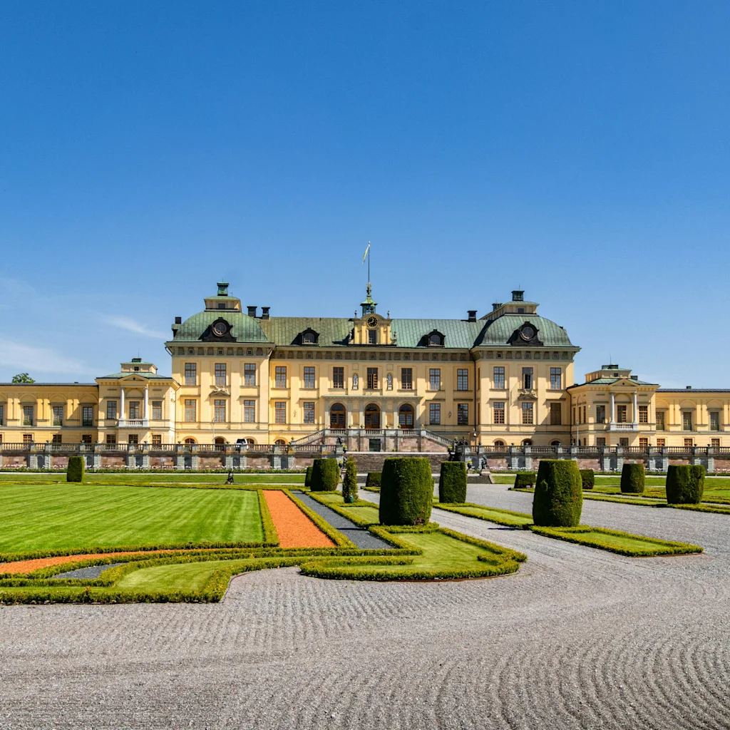Et storslået gult palads med grønne tage og kupler, omgivet af en formel have med grusstier og velplejede hække.