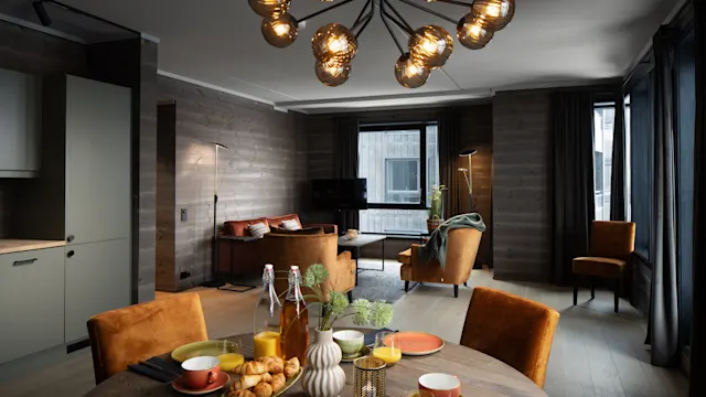 Dining table set for breakfast with croissants, juice, and colorful dishes; round wooden table in a modern living room with brown seating, large windows, and decorative overhead lighting.