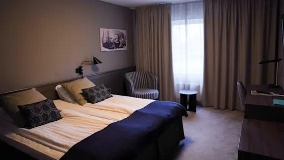 A tidy hotel room with twin beds, softly lit with wall sconces and overhead lighting, featuring a desk, chair, and framed wall art.