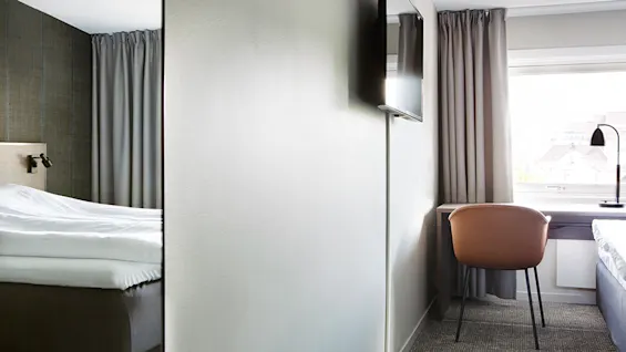 En seng står pænt i et moderne hotelværelse med et vægmonteret TV overfor. Et skrivebord og en stol er placeret ved siden af et stort vindue med lukkede gardiner.