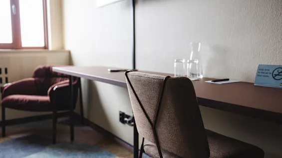 A chair faces a long desk holding glasses, a carafe, and a sign. A window and armchair are nearby. The sign reads, "Make your mark, take my card."
