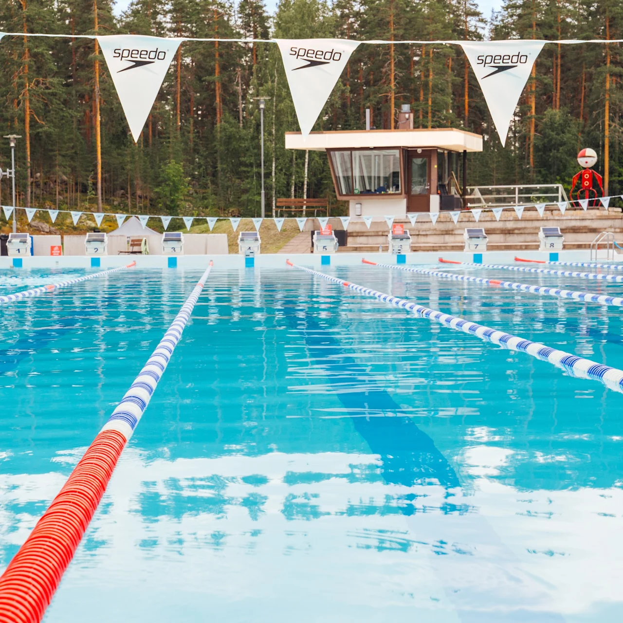 Outdoor swimming pool with clear lanes, surrounded by forest in the background. Bunting with "speedo" text hangs across the pool. Lifeguard tower visible with a person on duty.