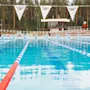 Ulkouima-allas selkeine ratoineen, ympärillä metsää taustalla. "Speedo" teksti liput roikkuvat altaan yllä. Uimavalvojan torni näkyvissä ja valvoja paikalla.