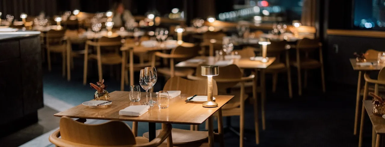 Bord som är elegant dukade med glas och servetter, upplysta av små lampor, är arrangerade i en dämpad, exklusiv restaurang. Trästolar omger varje bord, vilket förstärker den mysiga stämningen.