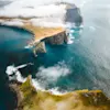 Cliffs rise steeply from the ocean, surrounded by swirling clouds and vibrant green grass. The sea crashes against the rocky shore, creating a dramatic coastal landscape.