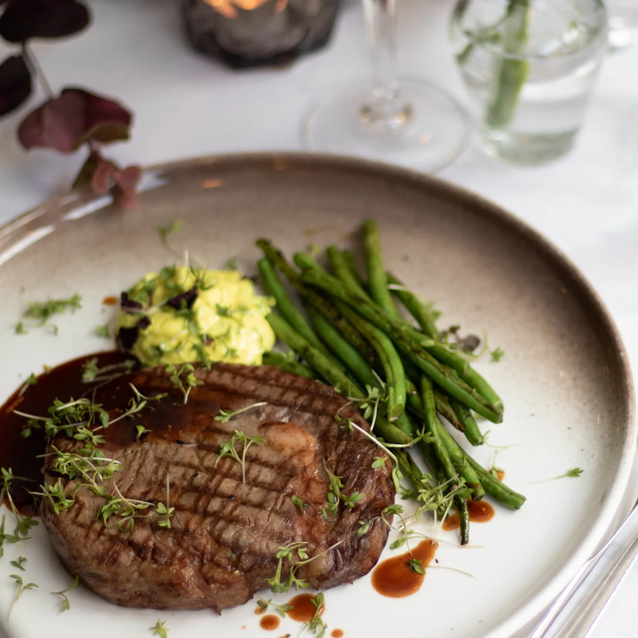 En biff toppet med spirer følger med grønne bønner og kartoffelmos på en keramiktallerken. En gaffel og kniv hviler på et marmorbord ved siden af et glas vand.