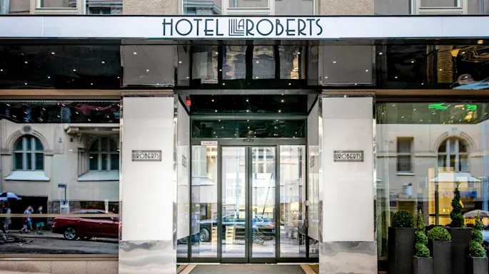 Inngang til Hotel Lilla Roberts med glassdører, som reflekterer den omkringliggende gaten. Skilt leser "Hotel Lilla Roberts", "ROOBERTS", og "PÄÄOVI MAIN ENTRANCE". Potteplanter rammer inn inngangen.