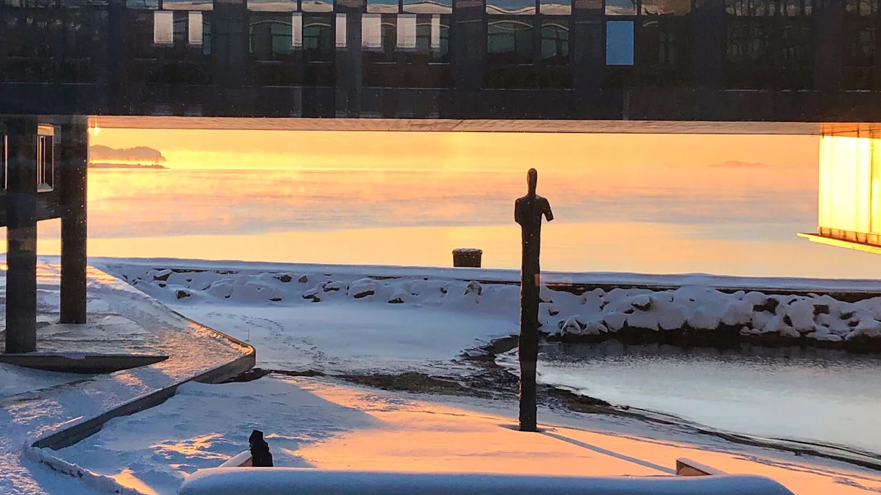 Lumisessa rantamaisemassa auringonlaskun aikaan patsas, heijastuksia rakennuksesta ja hehkuva valaisin.